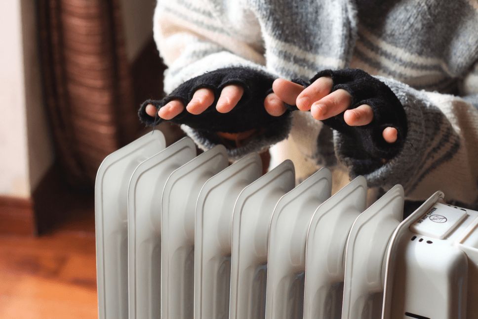 Person wearing a sweater and fingerless gloves warming hands over a radiator heater.
