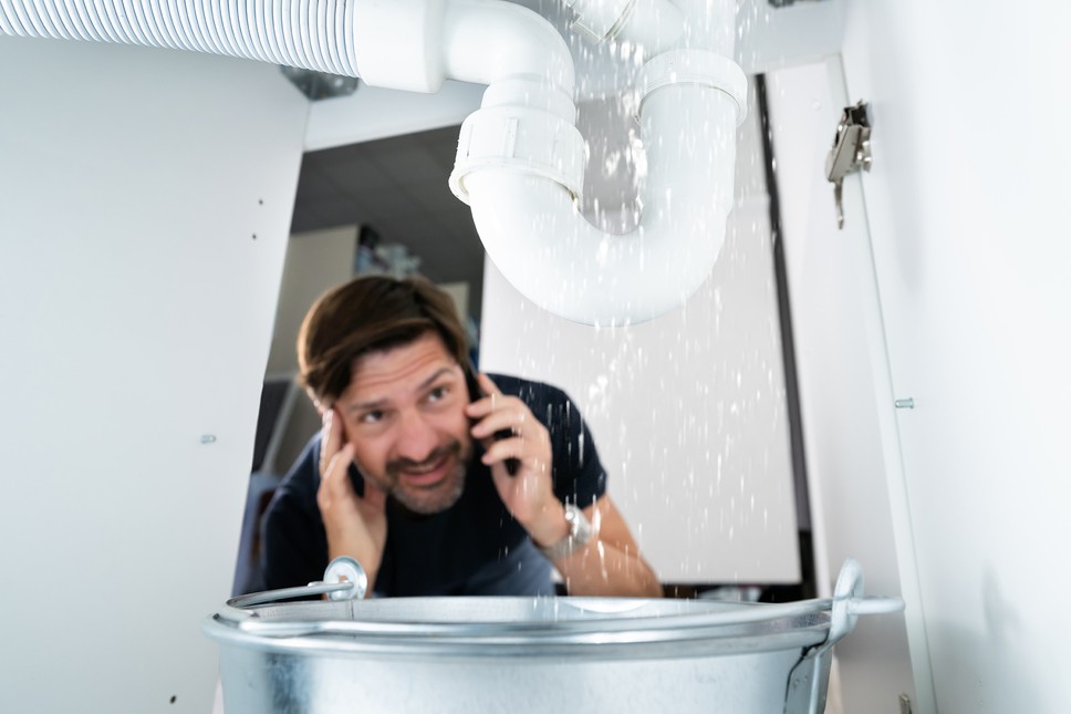An alarmed man on the phone looking under the sink at a pipe leaking rapidly into a bucket.